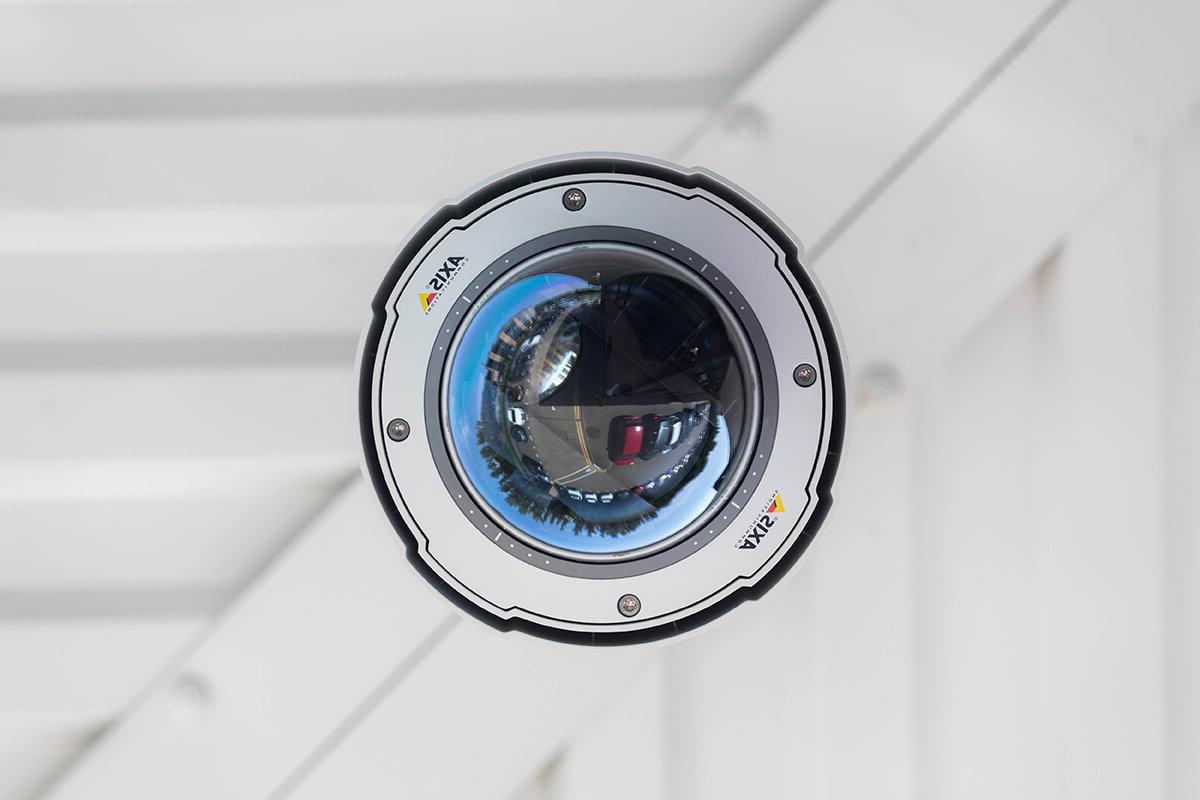 A close-up of a security camera against a white wall. The camera is Axis branded and the reflection of a car park can be seen in the camera